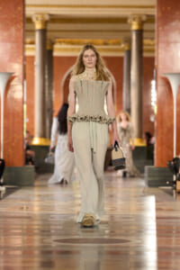 Model on a fashion runway wearing a beige pleated top with a sculpted ruffled peplum and wide trousers, carrying a small handbag.