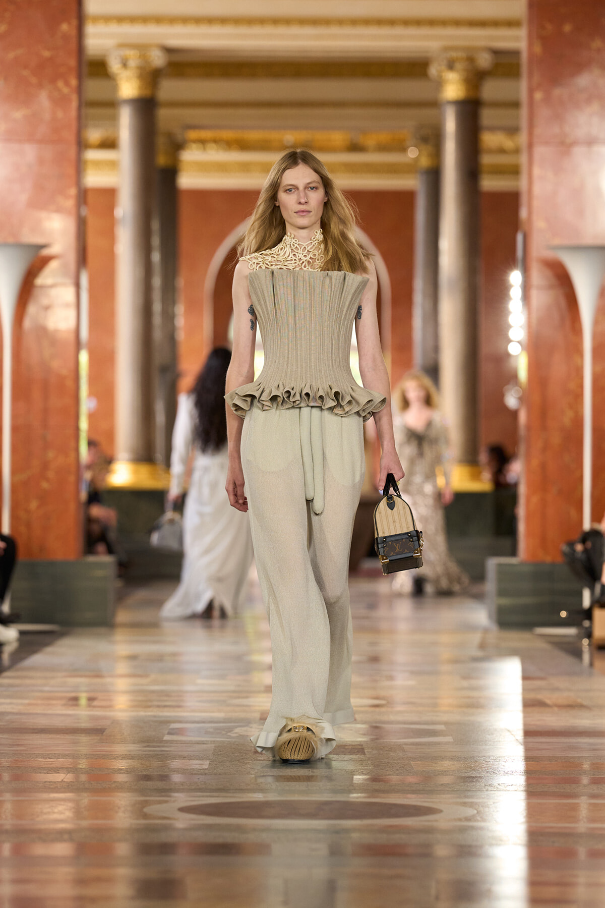 Model on a fashion runway wearing a beige pleated top with a sculpted ruffled peplum and wide trousers, carrying a small handbag.