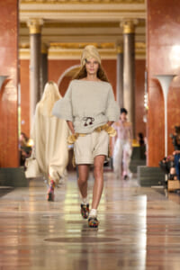 Model walks a runway in a beige knit sweater with oversized sleeves, matching shorts, tan turban, and bold patterned shoes in a grand, columned venue.