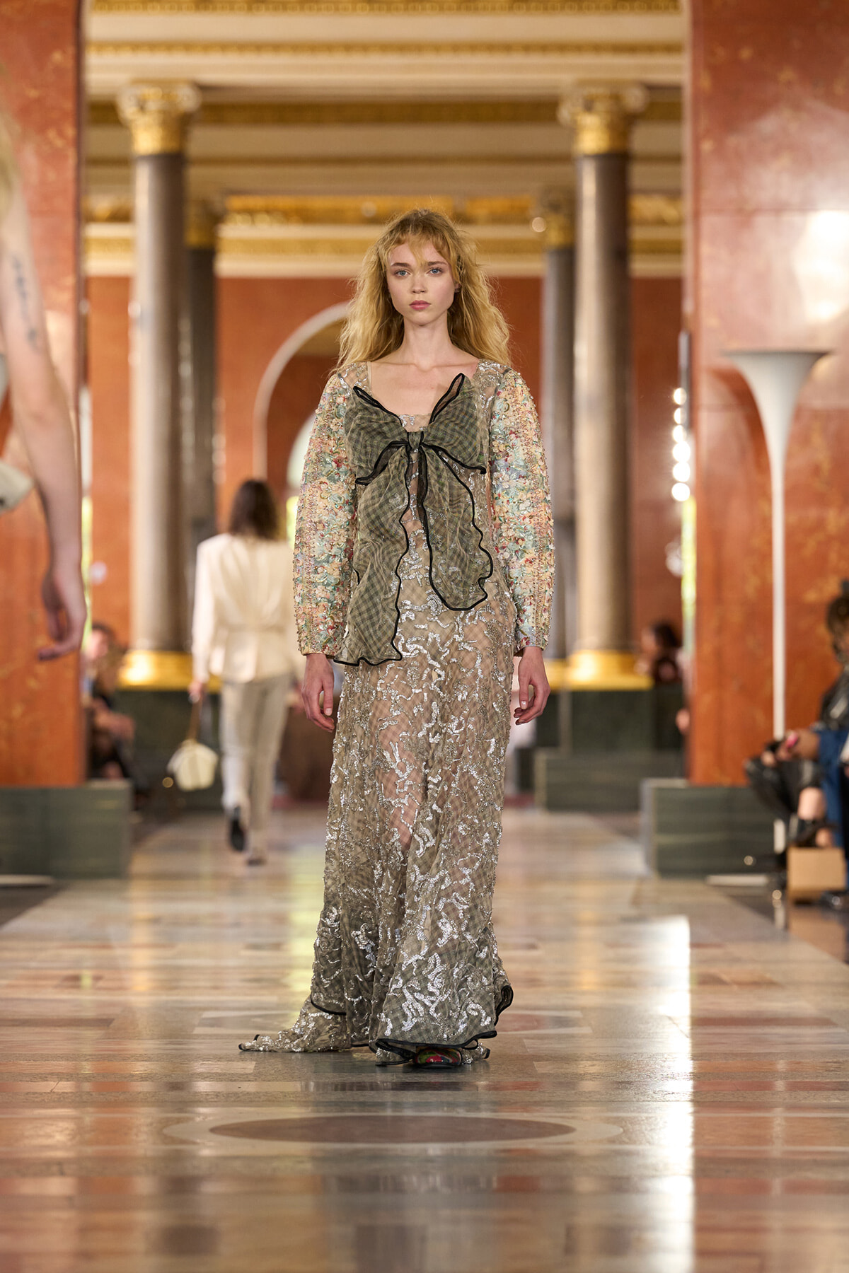 Model walks a marble runway in a floor-length silver lace gown with sheer floral sleeves and a dark green plaid bow at the bodice.