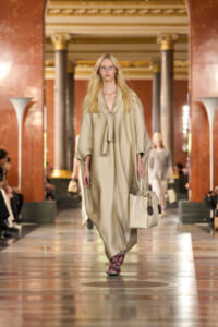Blonde model walks a runway in a beige flowing cape dress, carrying a light beige handbag inside an ornate marble hall.