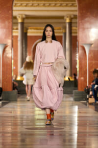 Model walks a fashion runway in a pale pink two-piece outfit with a voluminous skirt, carrying a fluffy beige bag.
