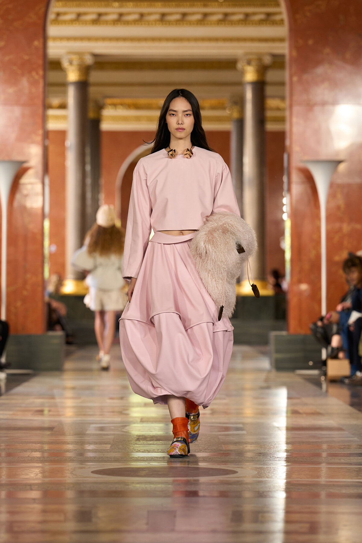 Model walks a fashion runway in a pale pink two-piece outfit with a voluminous skirt, carrying a fluffy beige bag.