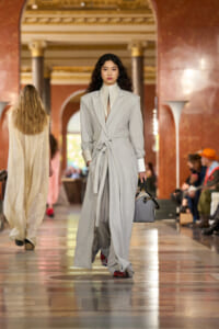 Model walking a runway in a light gray belted coat over a high-neck outfit, carrying a gray handbag.