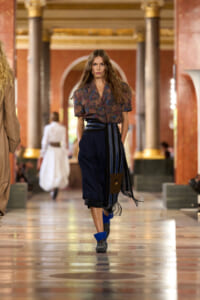 Female model walking a fashion show down a grand hall with red marble columns, wearing a patterned blouse and navy wide-leg cropped pants with a striped belt scarf.