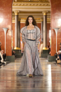 Model walks a high-fashion runway in a grand red-and-gold hall, wearing a sheer gray corseted dress with voluminous, striped sleeves and a flowing skirt, gold shoes visible at the hem.