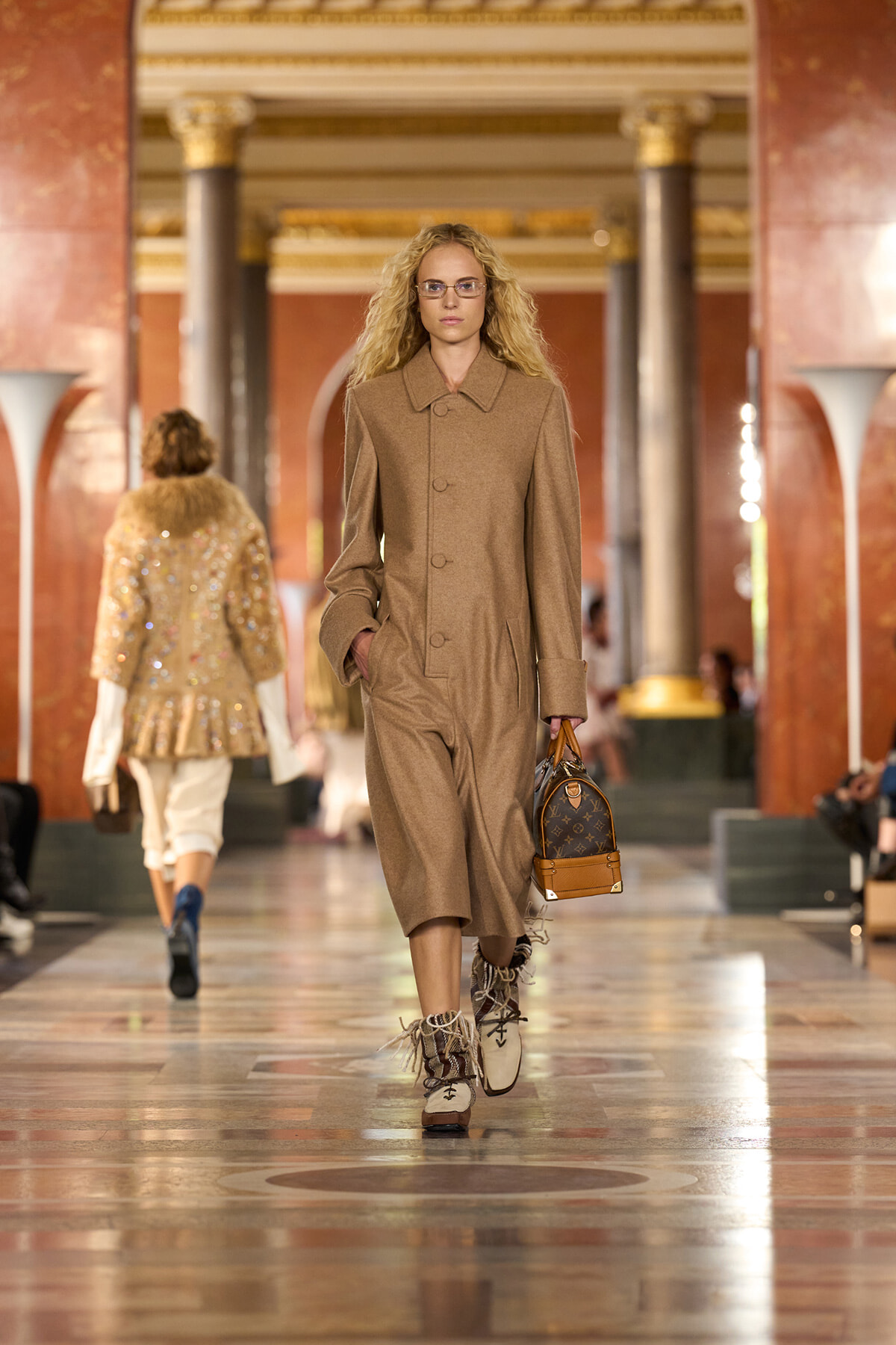 Model in a tan, buttoned coat walks a runway while carrying a brown designer handbag in a grand, marble-filled hall.