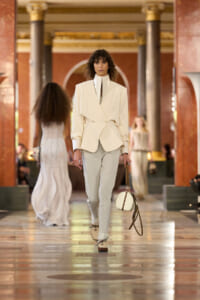 Model on a runway wearing a cream blazer with a high collar and light gray pants, carrying a round white bag with brown straps in an ornate hall setting.