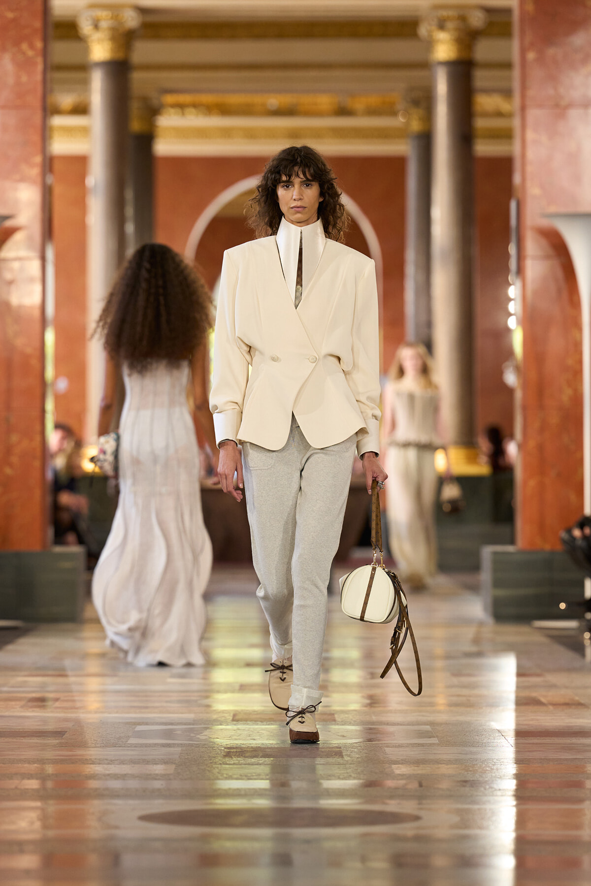 Model on a runway wearing a cream blazer with a high collar and light gray pants, carrying a round white bag with brown straps in an ornate hall setting.