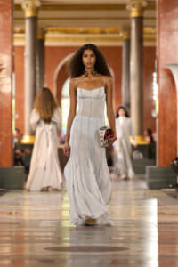 Model struts a light grey, strappy gown down a grand marble runway, clutch in hand.