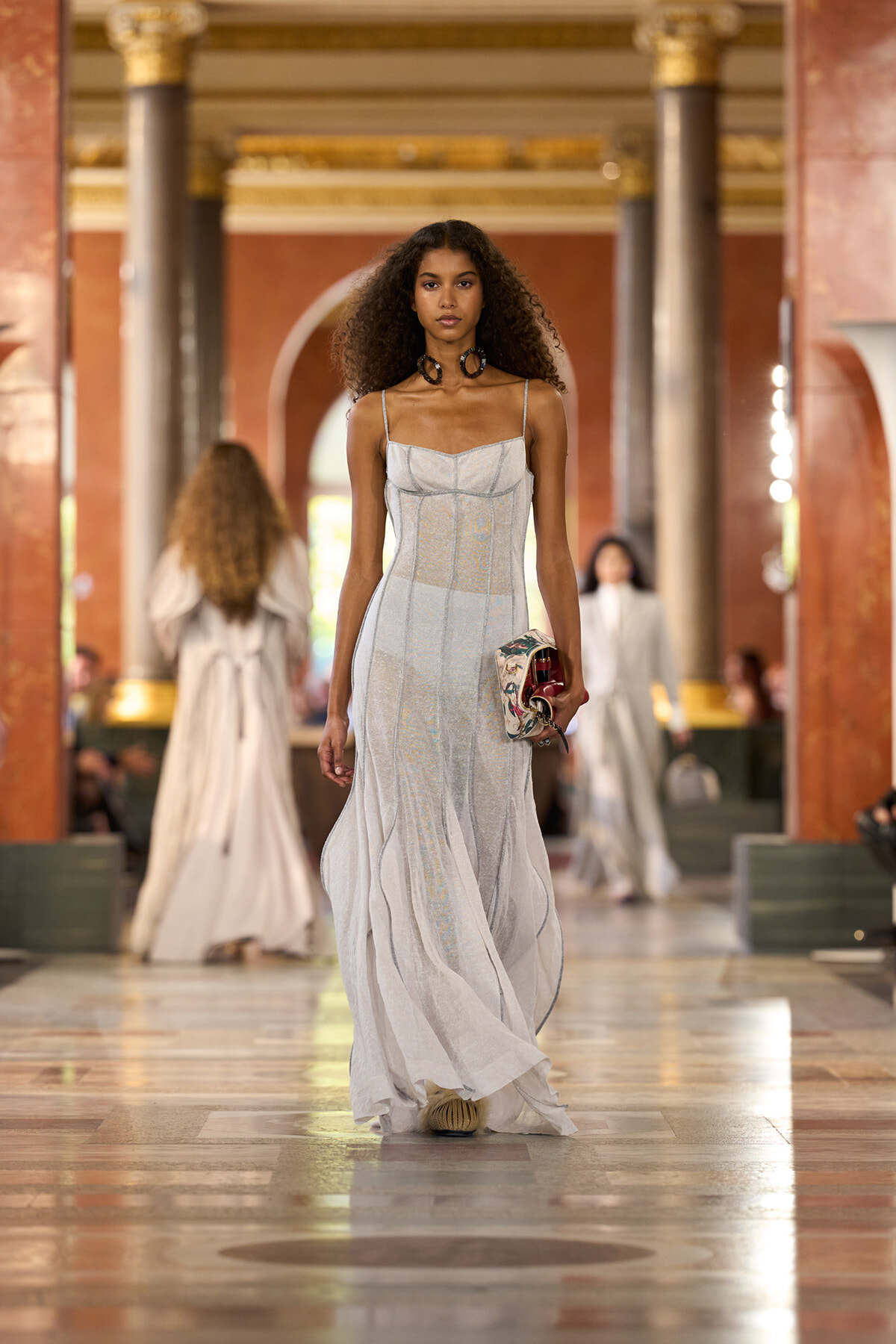 Model struts a light grey, strappy gown down a grand marble runway, clutch in hand.
