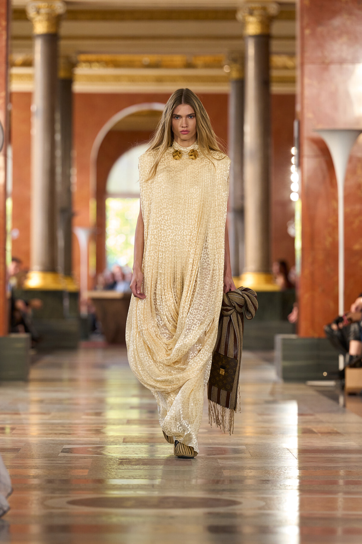 Model walks a runway in a floor-length cream lace dress with a draped silhouette, holding a striped scarf as she poses.