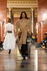 Runway model wearing a camel-textured coat with a tied belt and circular leather accents, carrying a patterned bag in a grand marble hall.