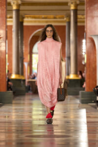 Model walking a fashion show in a pink lace high-neck dress, carrying a brown Louis Vuitton handbag in a grand marble hall.