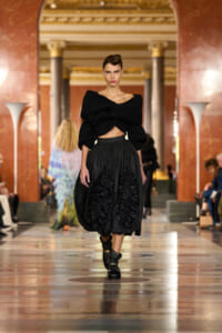 Model walking a runway in a black, oversized knit crop top and a full black skirt with ruched panels, in an ornate grand hall backdrop.