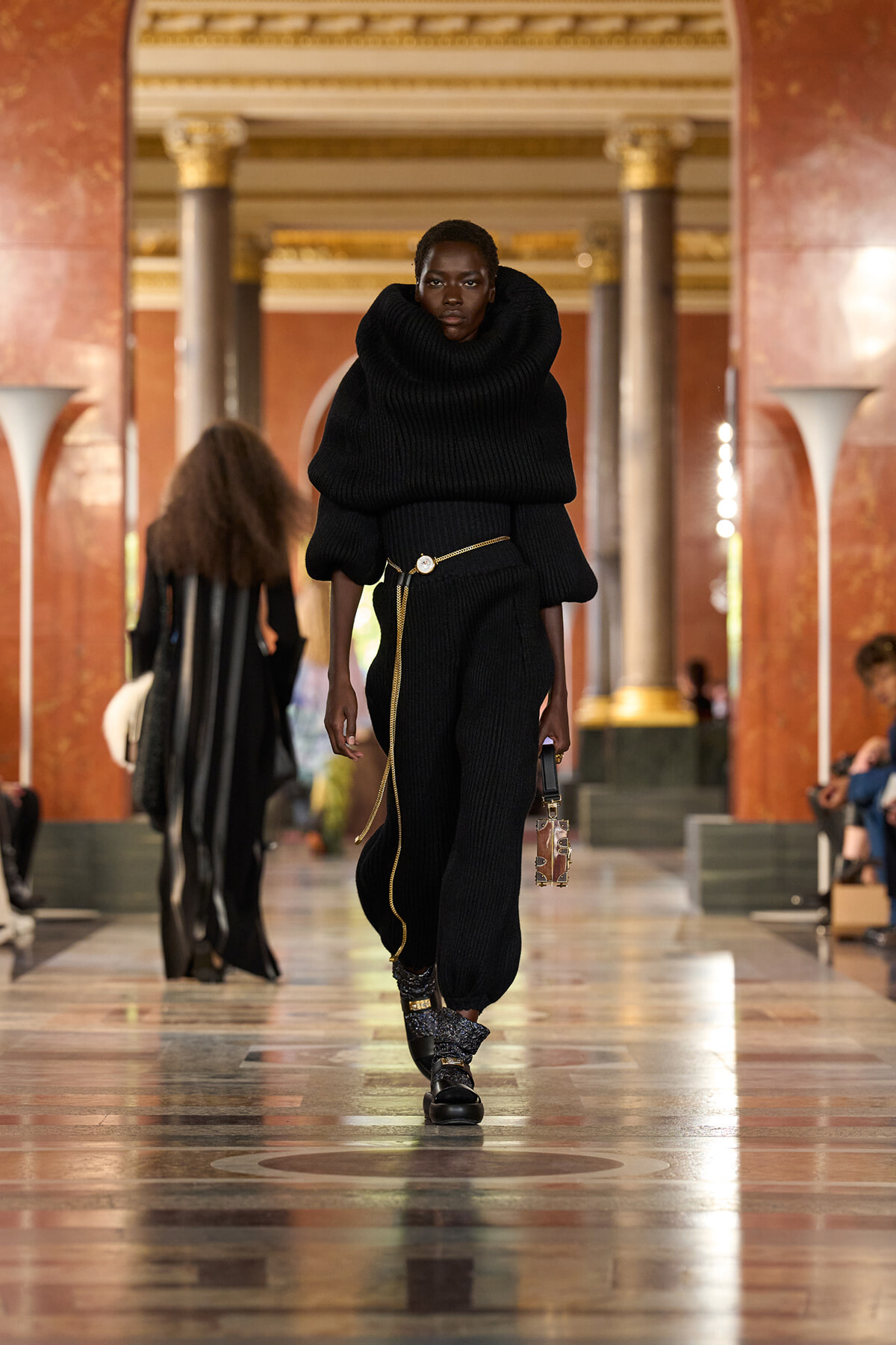 Model walks a runway in a black oversized knit sweater with a tall, sculptural collar and matching pants, cinched with a thin gold belt and a small handbag.