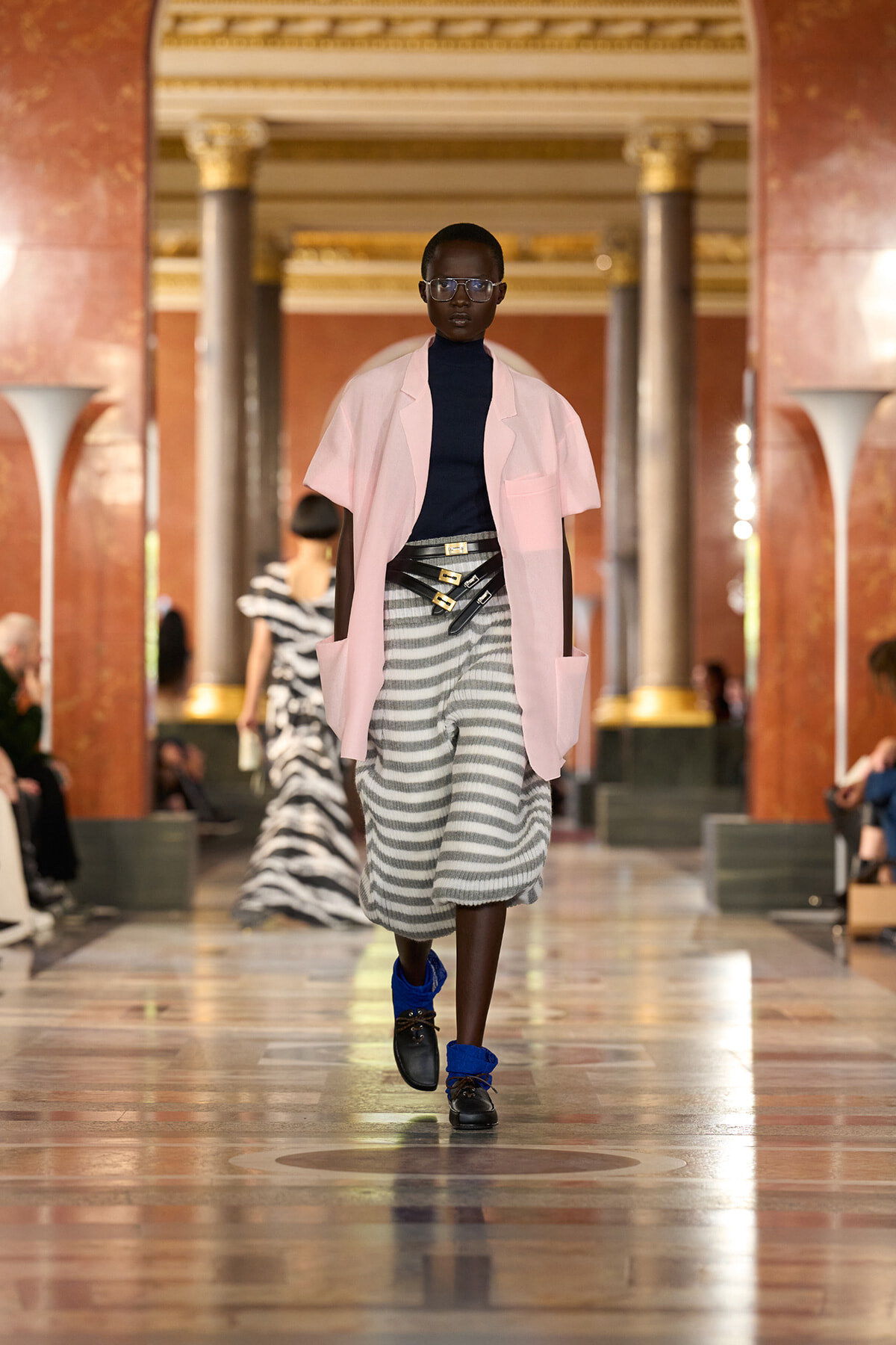 Runway model walks a grand hall in a pale pink oversized coat over a black turtleneck and striped gray skirt with a bold belt and blue socks.