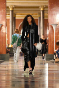 Model walks a fashion runway in a grand marble hall, wearing a black layered outfit with leather panels and a white fluffy bag.