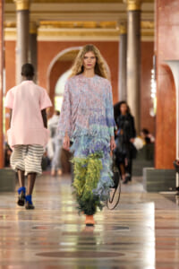 Model walks down a grand indoor runway in a pastel, multi-layered fringe dress with blue, green, and pink tones, carrying a small bag and wearing peach shoes against ornate architectural columns and arches.