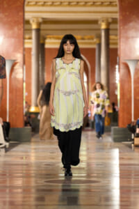 Model walking a fashion runway in a pale yellow, striped dress with floral trim over black pants inside an ornate marble hall with columns.