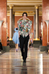 Model walks a runway in an opulent hall with marble columns, wearing a sculptural floral top over black pants and shiny shoes.