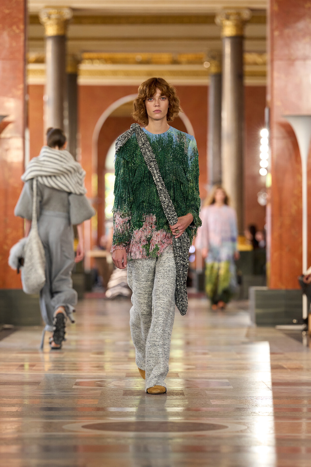 Model walking a fashion show in a grand marble hall, wearing a green-to-pink gradient knit top with fringe and gray pants, carrying a textured shoulder bag.