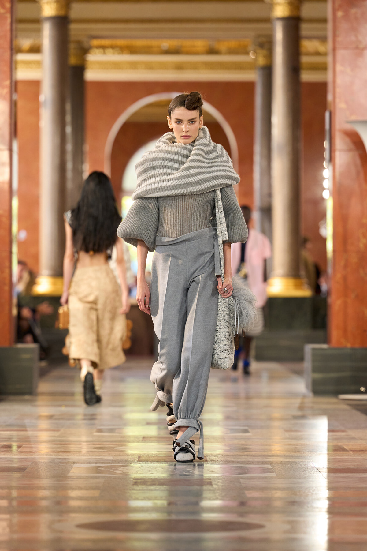 Model walking a fashion runway in a gray monochrome outfit: oversized striped scarf, chunky knit top, light gray tapered pants, and platform sandals, in a marble-hall venue with gold accents.