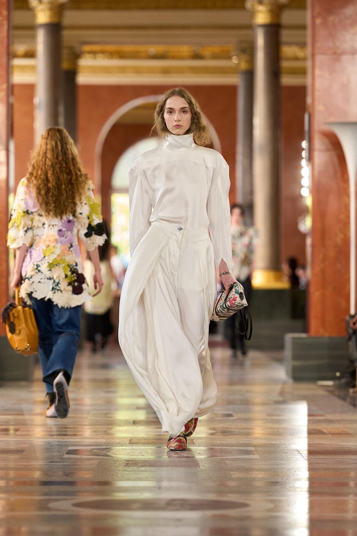 Female model walking down a marble hallway wearing an all-white high-neck ensemble and carrying a patterned clutch purse.