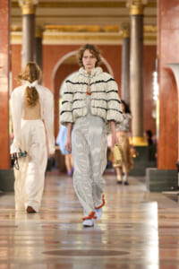 Model walks the runway in a striped jumpsuit with a voluminous cream quilted jacket edged in black, carrying a beige clutch in a grand hall setting.