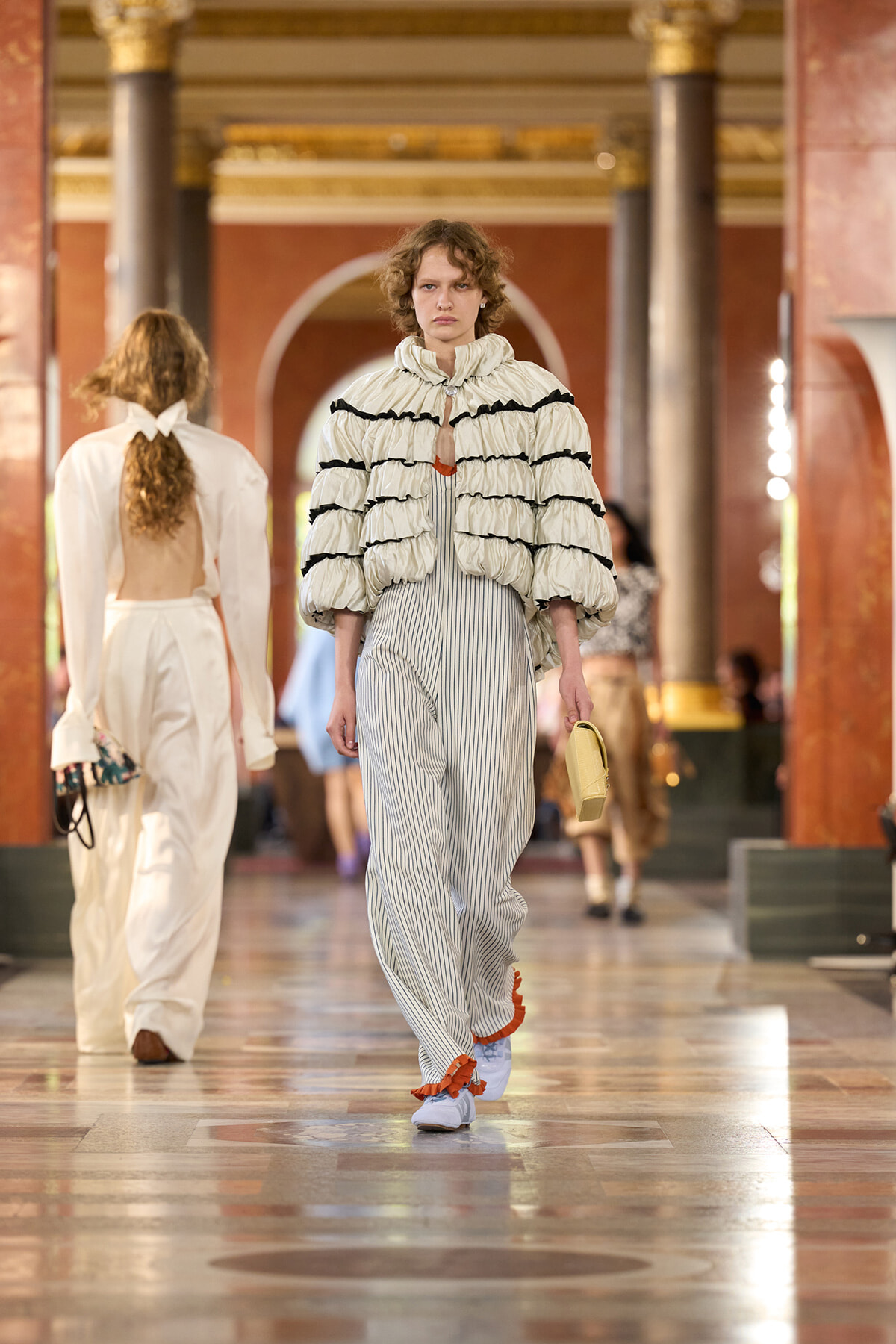 Model walks the runway in a striped jumpsuit with a voluminous cream quilted jacket edged in black, carrying a beige clutch in a grand hall setting.