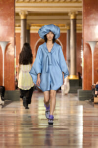 Model in a light blue, cape-style dress with a large matching hat walks a runway in a grand marble hall.