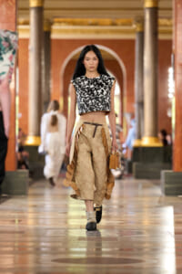 Model walking a fashion runway in a black-and-white brocade crop top and beige ornate trousers with circular side details, carrying a small tan bag.