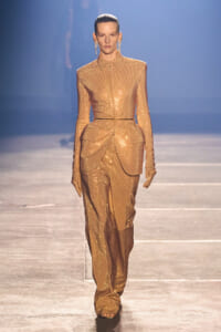 Model walks the runway in a glittering gold sequin suit with a structured peplum and long gloves.
