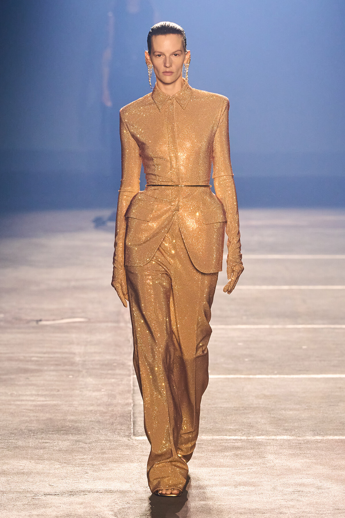 Model walks the runway in a glittering gold sequin suit with a structured peplum and long gloves.