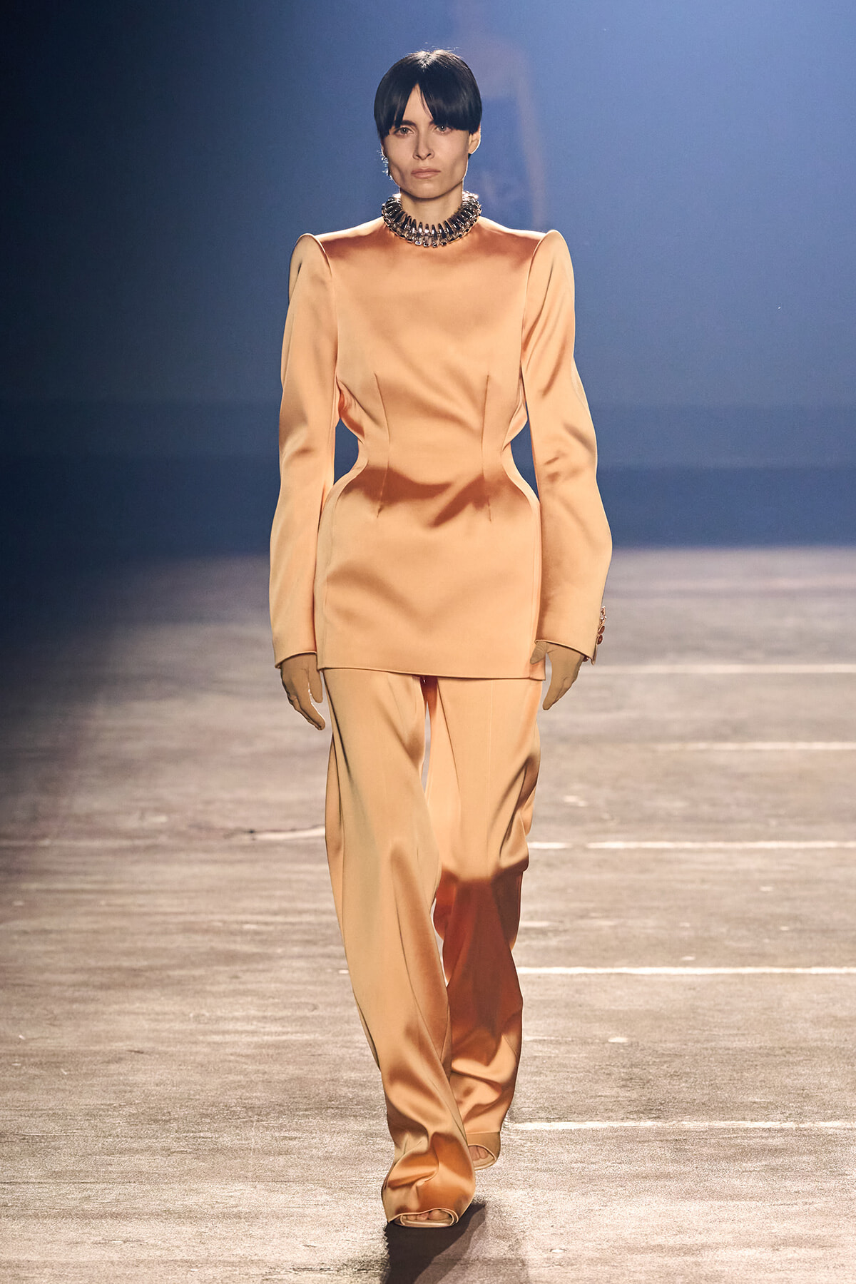 Model walking the runway in a peach satin tailored pantsuit with a sculpted top and statement choker necklace on a dim catwalk.