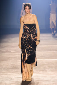 Model on a runway wearing a strapless black-bronze velvet gown with peach panels and long nude gloves.