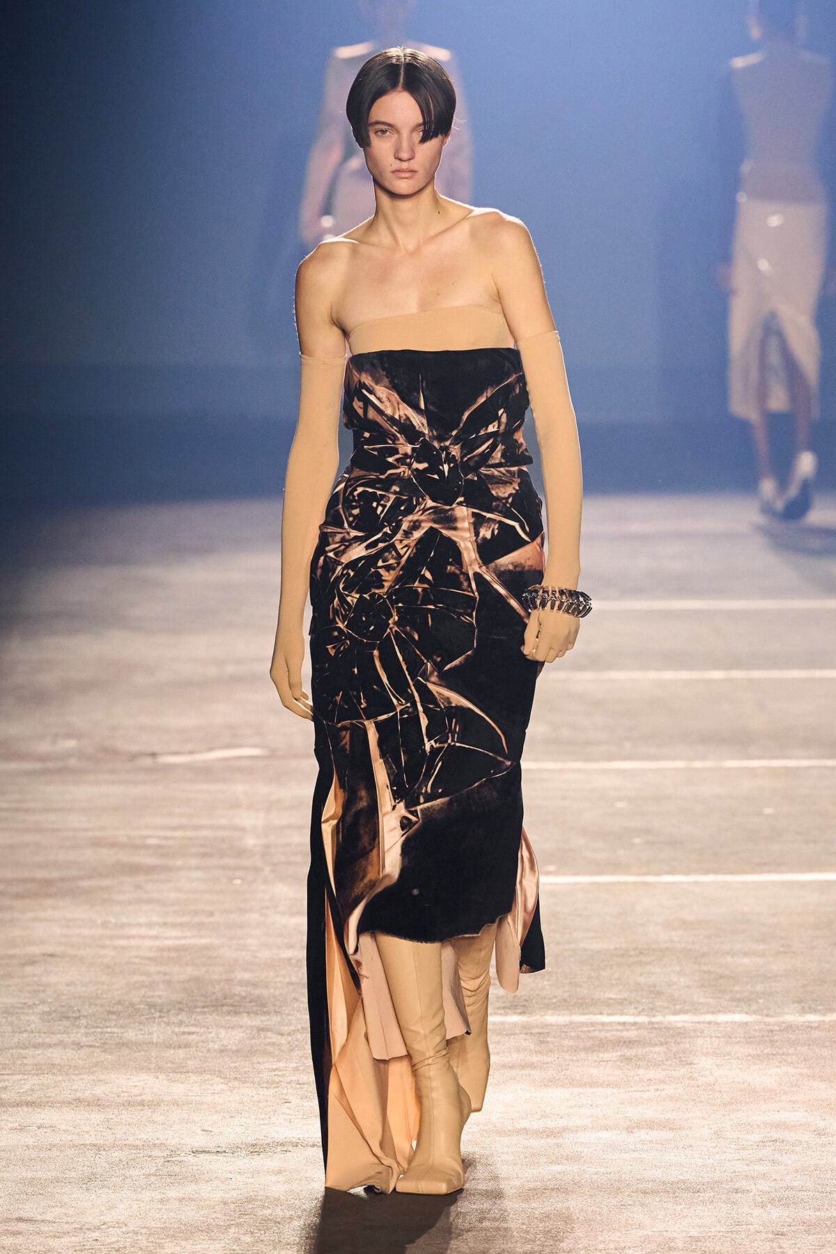 Model on a runway wearing a strapless black-bronze velvet gown with peach panels and long nude gloves.