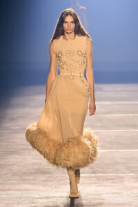 Model on a runway wearing a sleeveless nude-beige gown with black embroidery, a draped wrap skirt, and a voluminous feathered hem with beige boots.