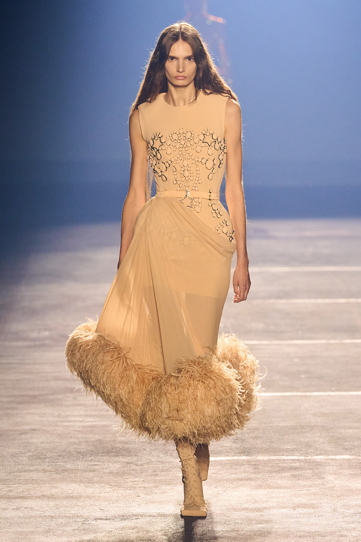 Model on a runway wearing a sleeveless nude-beige gown with black embroidery, a draped wrap skirt, and a voluminous feathered hem with beige boots.