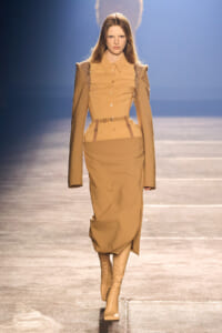 Model walking the runway in a camel-toned, coordinated outfit with structured shoulders and knee-high boots.