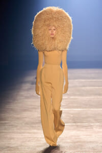 Runway model in a large fluffy beige headpiece and matching beige wide-leg trousers walks the catwalk.