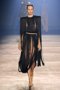 Model walking the runway in a black fringe dress with a beige underlayer and a slim belt, wearing statement earrings.
