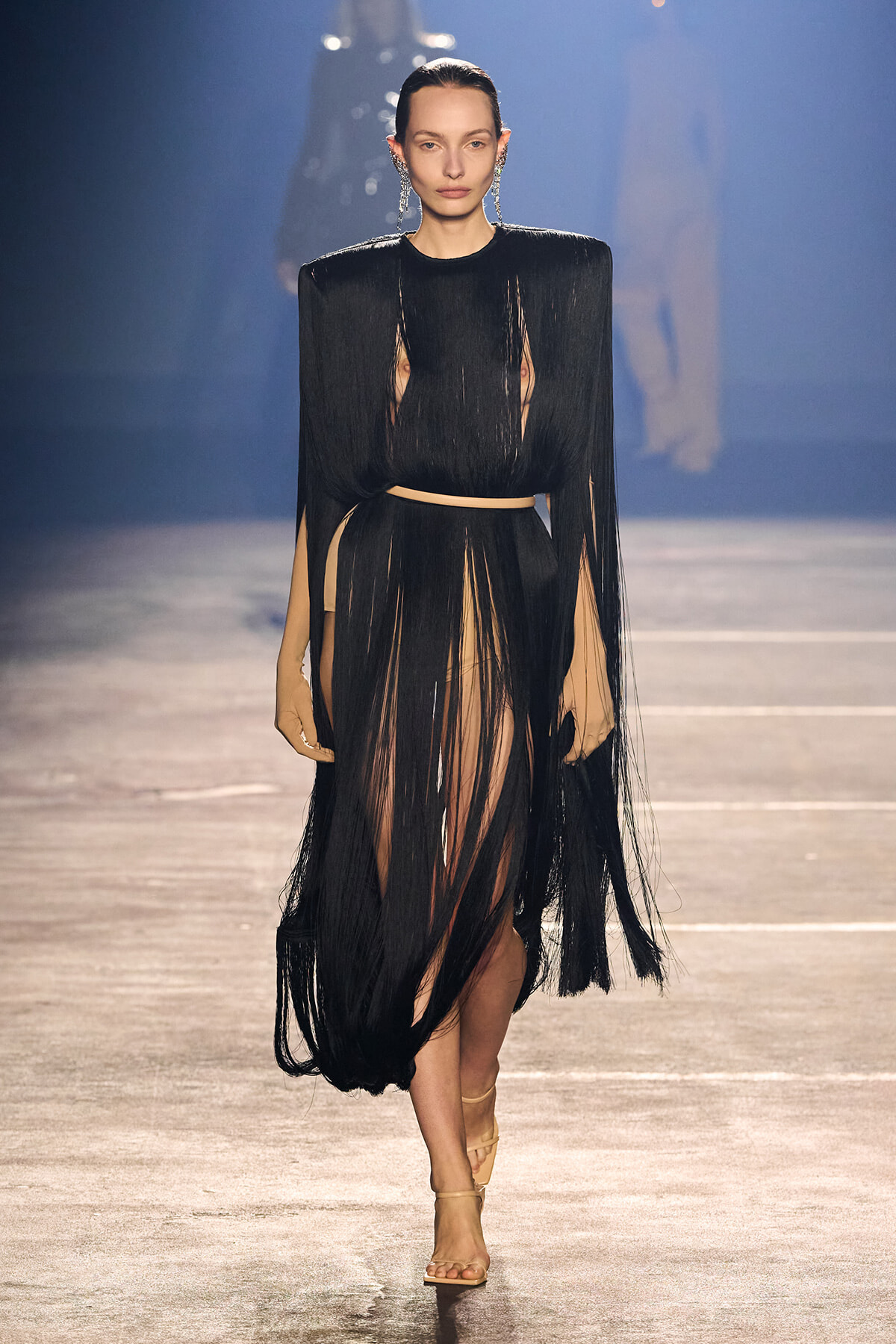 Model walking the runway in a black fringe dress with a beige underlayer and a slim belt, wearing statement earrings.