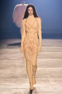 Model walking a beige bodysuit covered in cascading pearl bead chains on a fashion runway.