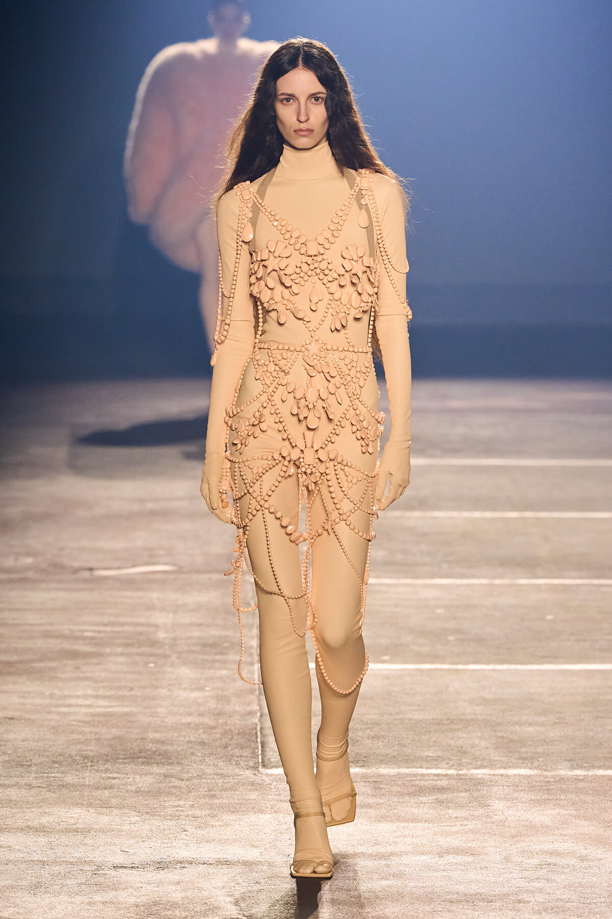 Model walking a beige bodysuit covered in cascading pearl bead chains on a fashion runway.