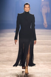 Model on a runway wearing a black tailored blazer with a long fringe skirt and strappy sandals, dark earrings visible under slicked-back hair