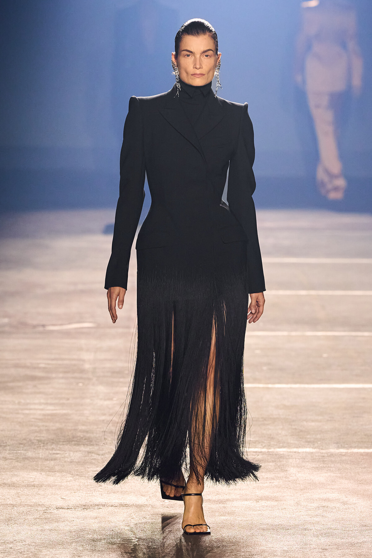 Model on a runway wearing a black tailored blazer with a long fringe skirt and strappy sandals, dark earrings visible under slicked-back hair