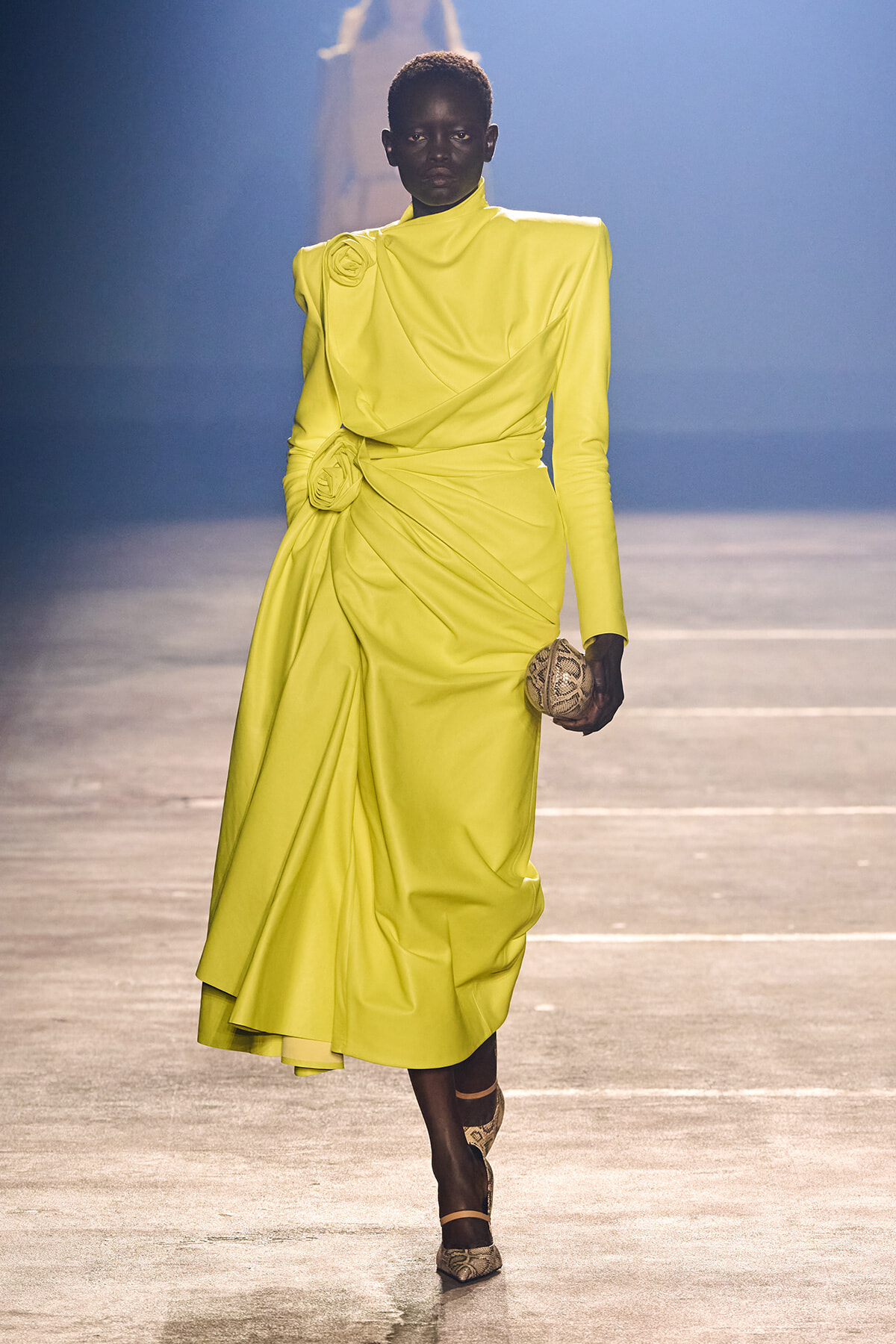 Model walking a runway in a neon-yellow draped gown with a floral rosette, holding a small patterned clutch.