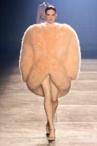 Model walks a fashion runway wearing a voluminous peach faux-fur coat that envelops her silhouette, paired with nude heels.