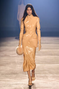 Model walking a runway in a peach patent leather suit with a long-sleeve jacket and matching midi skirt, carrying a round fluffy beige handbag.